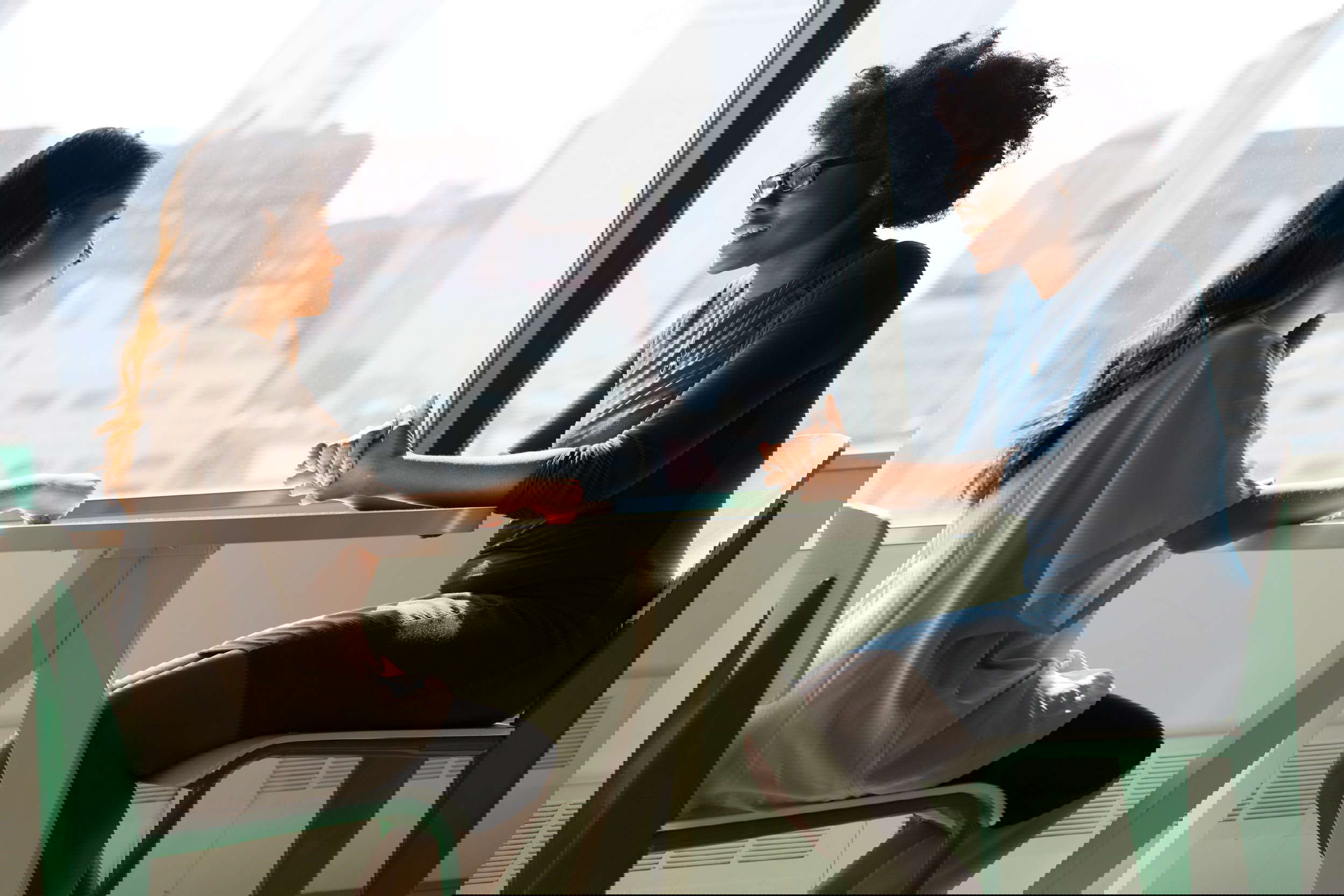 A group of four young women are sitting around a rectangular table in an indoor setting. All of them are wearing casual clothing. Two of them are sitting side by side at the table and appear to be talking to one another, one of them is smiling and gesturing with her hands. The other two are sitting across from each other, one of them is looking directly at the camera while the other is looking down at the table. All four of the women have their hands placed on the table in front of them. The table is made of light wood and is set against a wall with a patterned wallpaper.