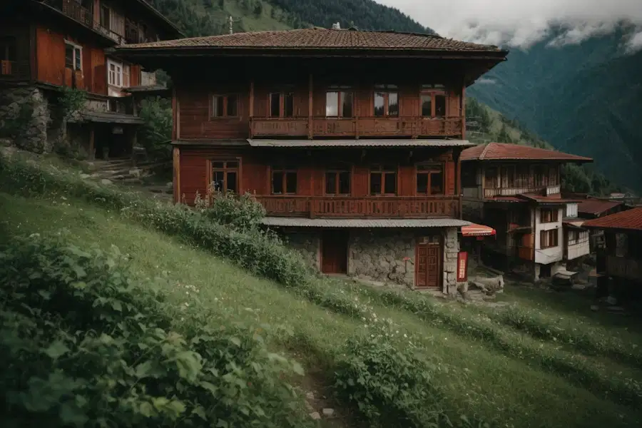 This image captures a picturesque scene of a building nestled on a verdant hill. The building, which appears to be a house, exudes a rustic charm with its stone walls, dark wooden door, and dormer window. A balcony is also noticeable, perhaps serving as a vantage point for the breathtaking surroundings. Nearby, another house with similar stone architecture can be seen. The foreground is lush with green grass and bushes, enhancing the overall rural feel of the scene. In the distance, a majestic mountain range capped with clouds looms, adding a sense of grandeur to the tranquil setting. The picture seems to be a perfect representation of serene village life, a retreat nestled in nature.